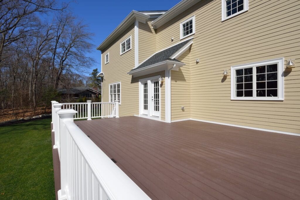 A spacious, brown wooden deck attached to a beige house with white trim, featuring white railings and double glass doors—perfect for relaxing or learning how to clean Trex decking—overlooking a grassy yard and trees under a clear blue sky.