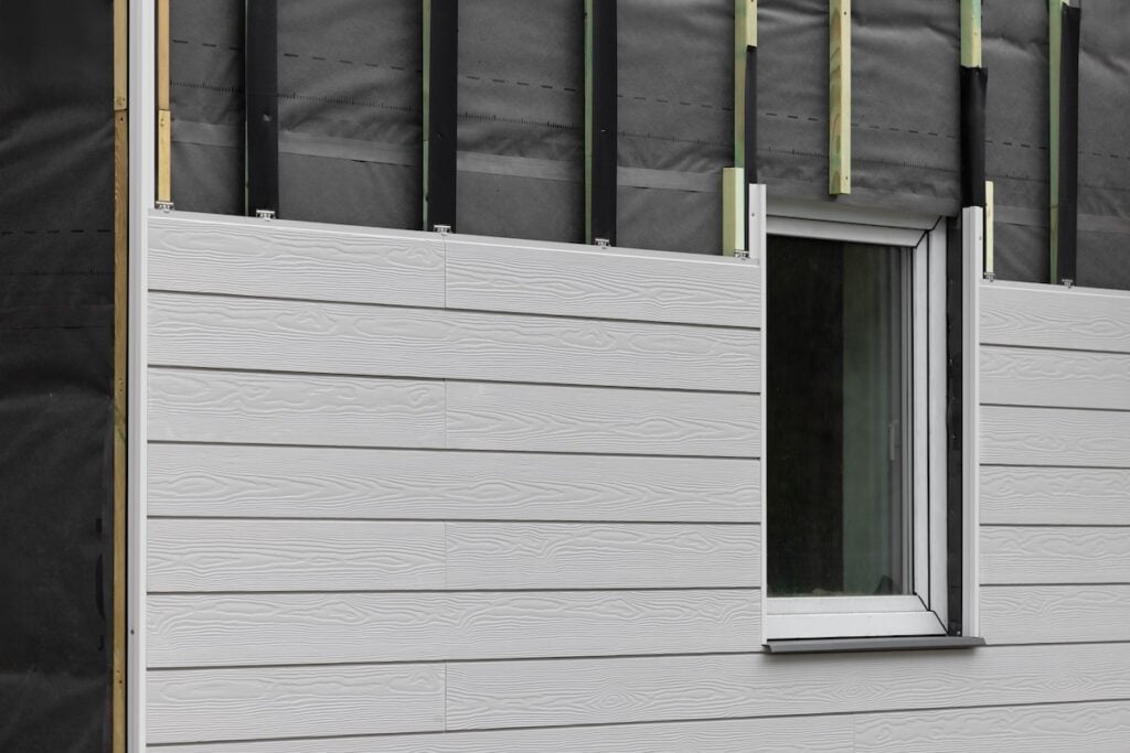 Partially installed light gray siding panels on a building wall around a window, with exposed black weather barrier and vertical furring strips visible above the siding.