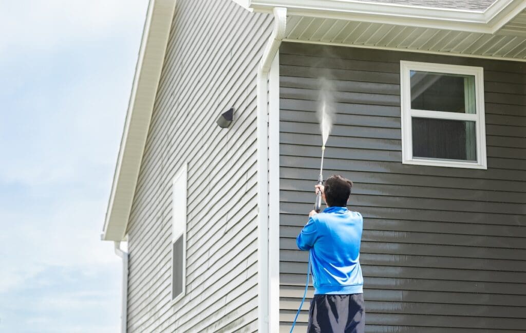 A person in a blue shirt uses a pressure washer to clean the siding of a gray house on a sunny day.