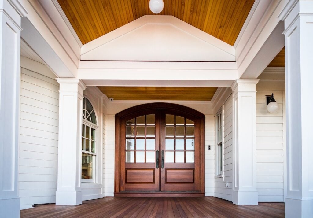 A spacious front porch with white columns, a wood-paneled ceiling, and double wooden doors with glass panes