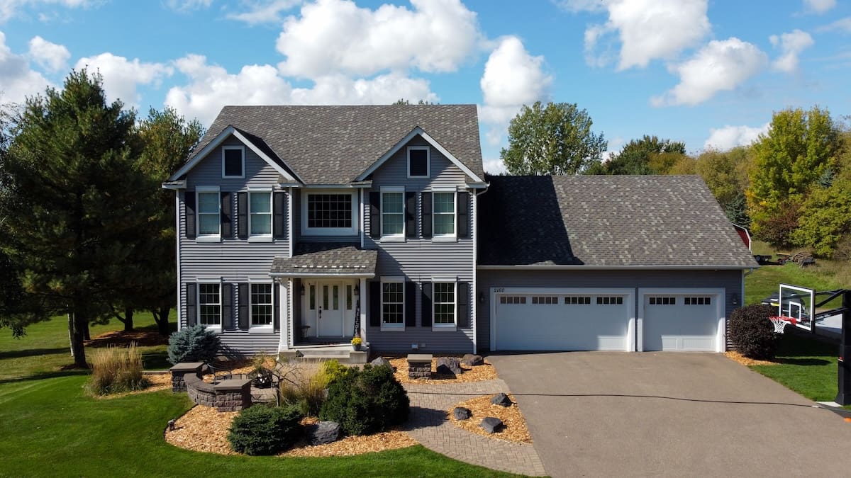 Two-story gray house with sustainable siding and white trim, three-car garage, and landscaped front yard featuring bushes, rocks, and mulch. Trees and a green lawn surround the property under a partly cloudy blue sky.