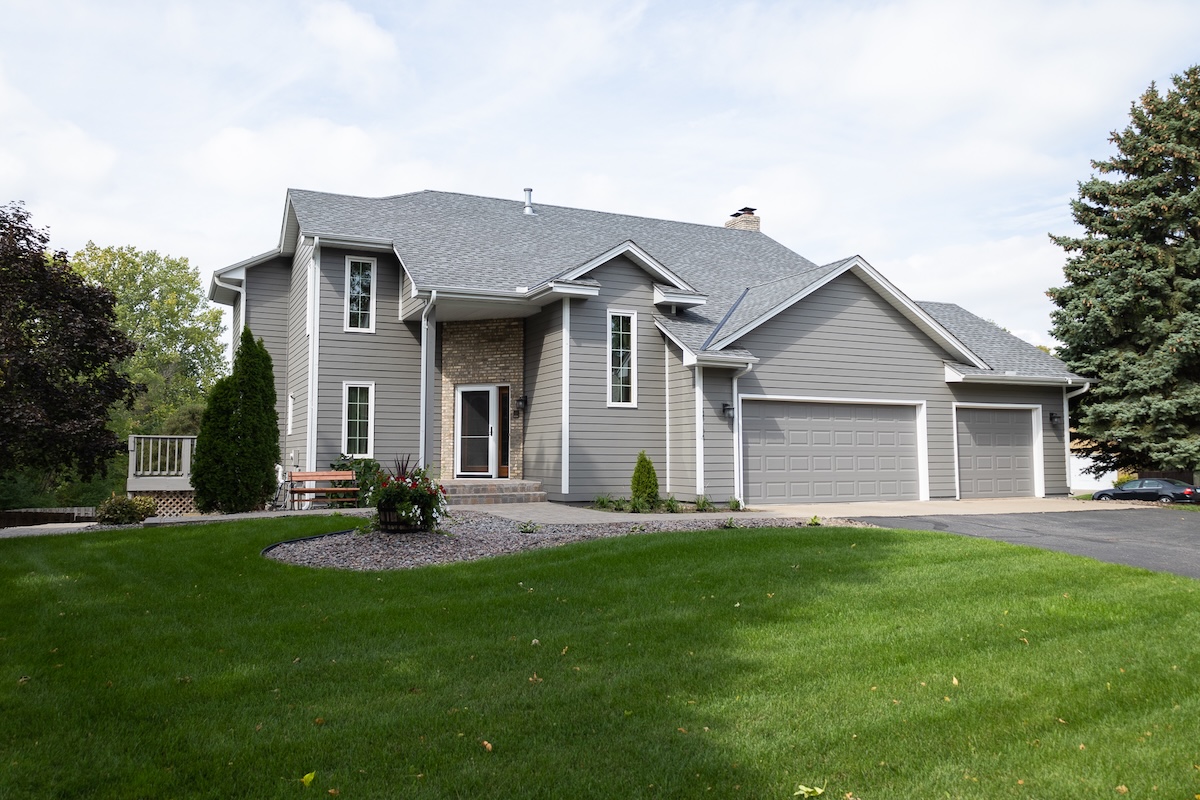 A modern two-story gray house with sustainable siding, a large driveway, three-car garage, front porch, and well-maintained lawn, surrounded by trees and shrubs on a clear day.