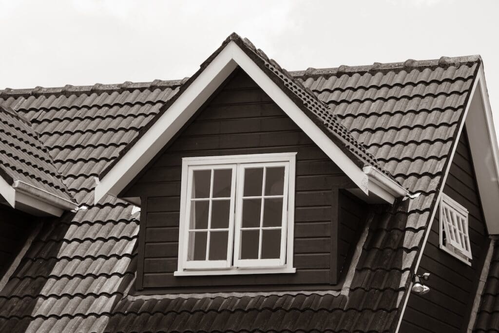 A sepia-toned photo of a house roof with dark tiles and a dormer window framed in white trim,