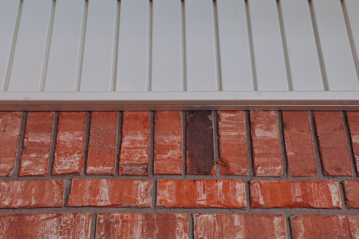 Close-up of a red brick wall featuring one dark brown brick among the red ones, topped by beige vinyl siding—showcasing unique vinyl siding and brick combinations.