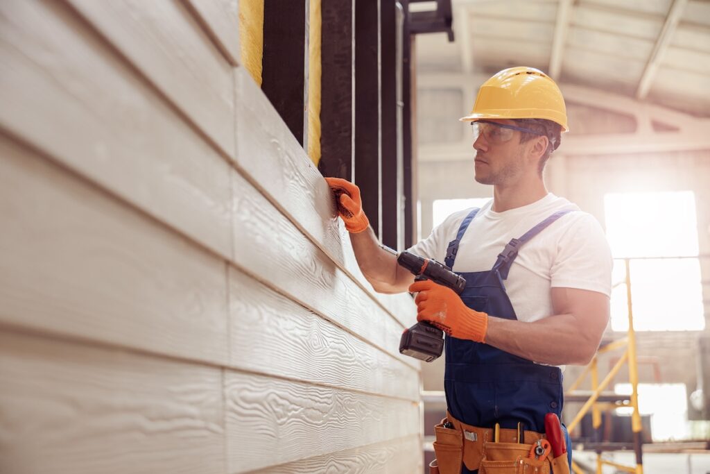 A james hardie certified installer in a hard hat, gloves, and overalls uses a power drill while installing or inspecting siding inside a well-lit industrial structure.