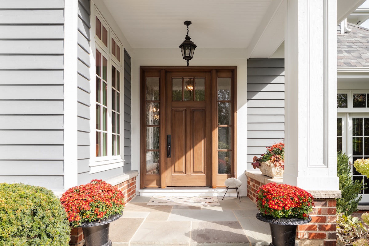 A front porch with a wooden door, sidelights, and a black lantern above. Potted red flowers decorate each side of the entrance, surrounded by gray most durable siding with white trim. Sunlight brightens the space.