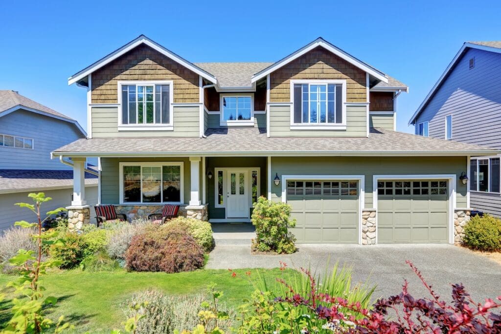 Two-story suburban house with green siding, embracing current siding color trends, stone accents, double garage doors, front porch with white pillars, and a landscaped yard with shrubs and flowers under a clear blue sky.