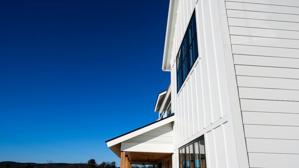 A modern white house with large windows is shown against a bright blue sky, expertly finished by James Hardie certified installers. Minimal landscaping is visible, with a hint of distant trees on the horizon.
