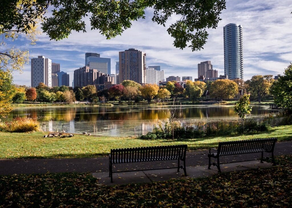 Two empty park benches face a calm pond, with autumn trees and city skyscrapers in the background. Fallen leaves cover the ground, branches frame the top—capturing the peaceful side of things to do in the Twin Cities.