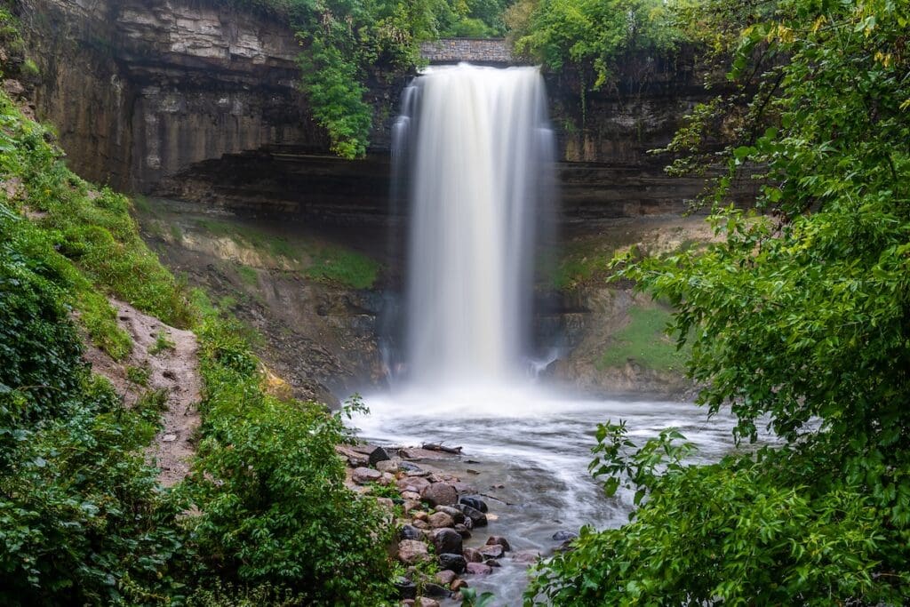 A tall waterfall (Minnehaha Falls) cascades over a rocky cliff into a pool below, surrounded by lush green trees and foliage under a cloudy sky—a breathtaking highlight among things to do in the Twin Cities.
