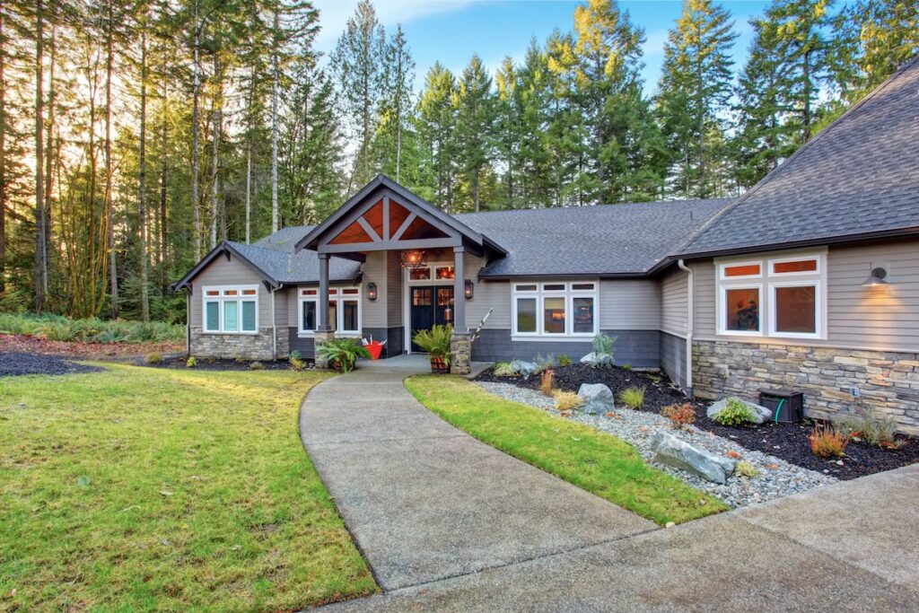 Single-story modern home with sustainable siding, stone accents, and large windows, nestled among tall trees. A curved concrete walkway leads to the covered entryway. The front yard is landscaped with grass, rocks, and mulch.