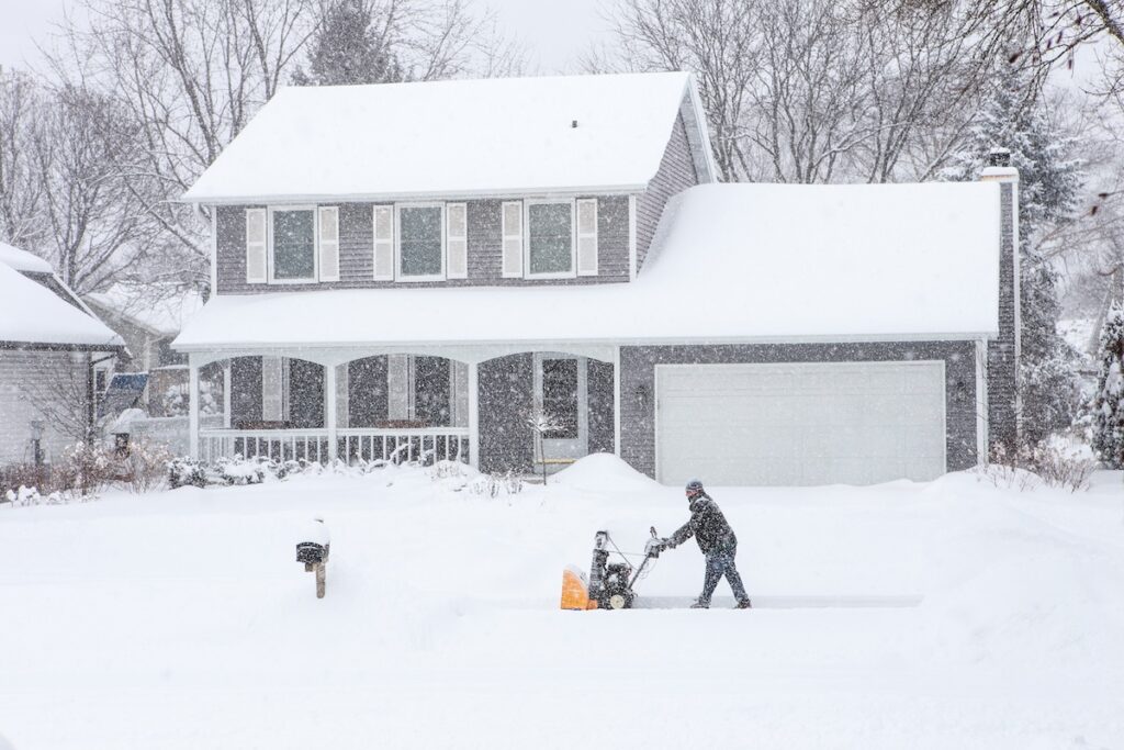 A person uses a snow blower to clear a driveway in front of a house with weatherproof siding, surrounded by snow-covered trees and rooftops during a heavy snowfall in a suburban neighborhood.