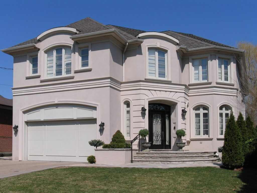 Two-story suburban house with light beige stucco exterior, one of the most durable siding options, arched windows, double garage, and decorative front entrance with black door, steps, potted plants, and outdoor lanterns against a clear blue sky.