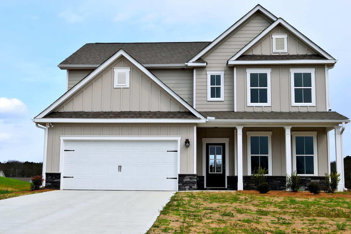 A modern two-story suburban house with sustainable siding in beige, dark grey roof, white trim, a double garage, covered front porch, and a concrete driveway, with a lawn and small shrubs in front.