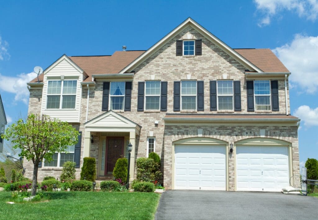 A two-story suburban house with a beige brick exterior accented by vinyl siding and brick combinations, black shutters, double garage doors, and a well-kept lawn with shrubs and a small tree under a blue sky with some clouds.
