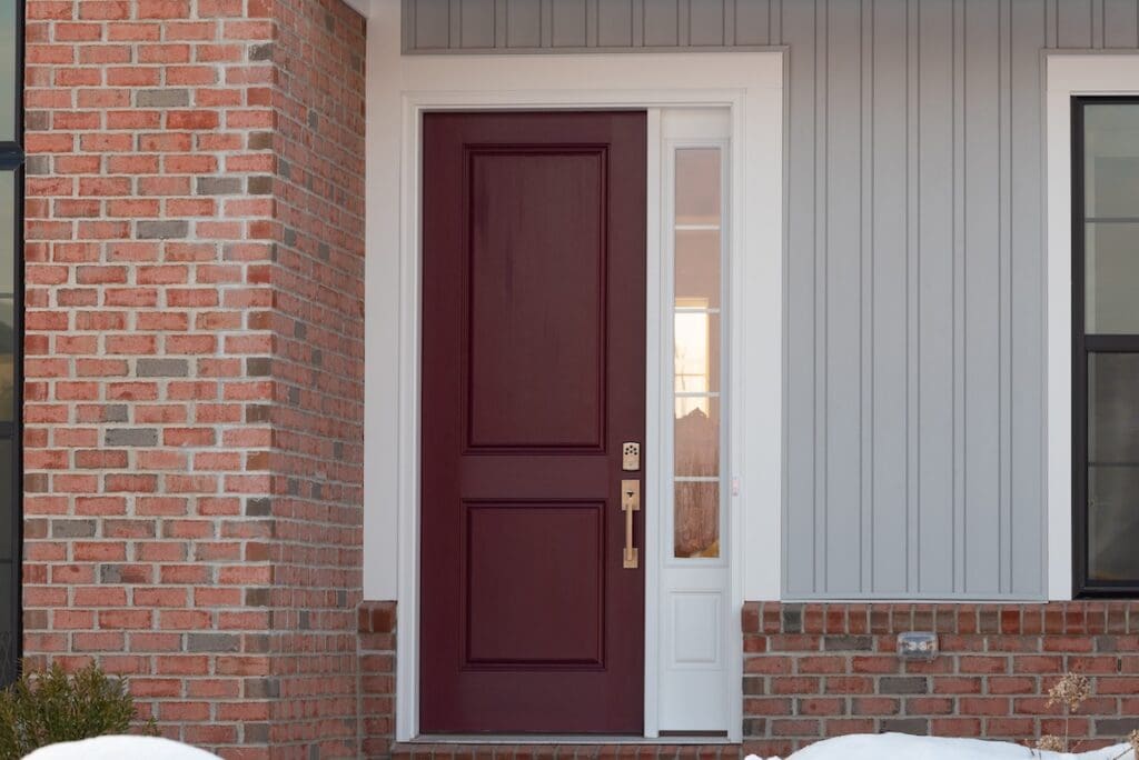 A maroon front door with a glass side panel and gold hardware stands out on a house exterior showcasing vinyl siding and brick combinations