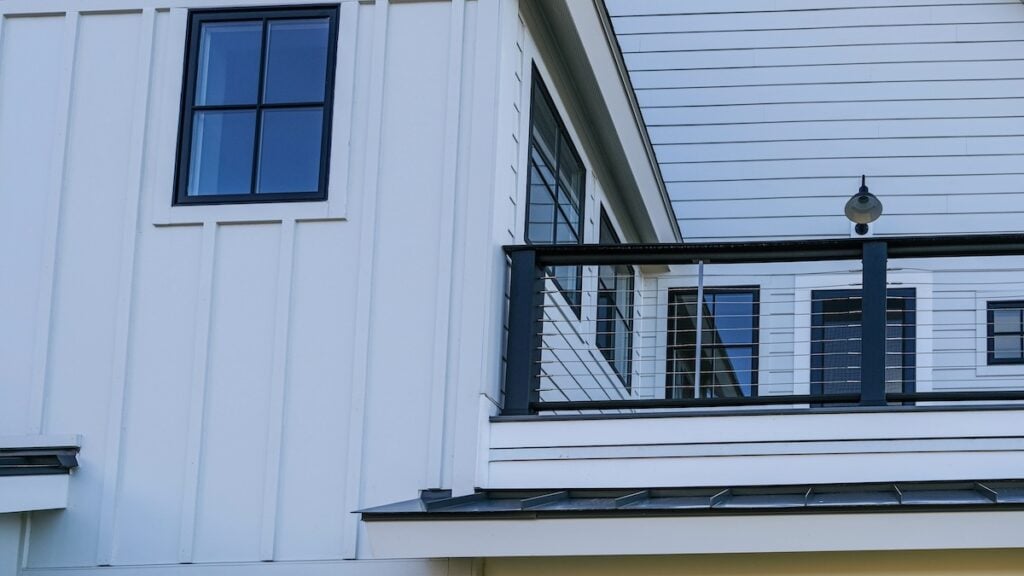 Modern house exterior featuring the most durable siding in crisp white vertical panels, black-framed windows, and a balcony with metal cable railing and a wall-mounted outdoor light fixture.