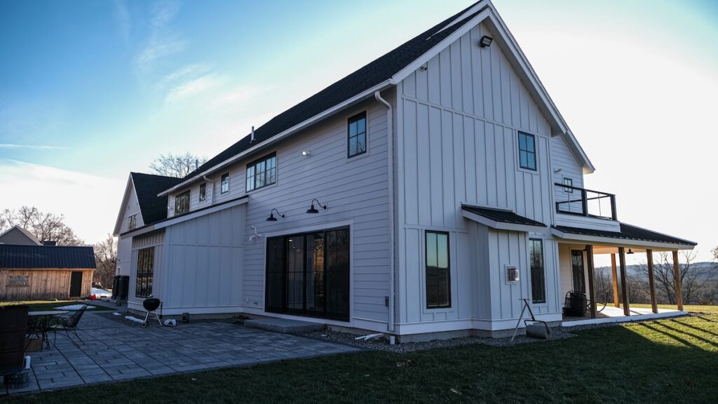 Modern two-story farmhouse with white board-and-batten siding installed by James Hardie certified installers, black framed windows and doors, and a spacious patio in the backyard, set against a clear sky and open landscape.