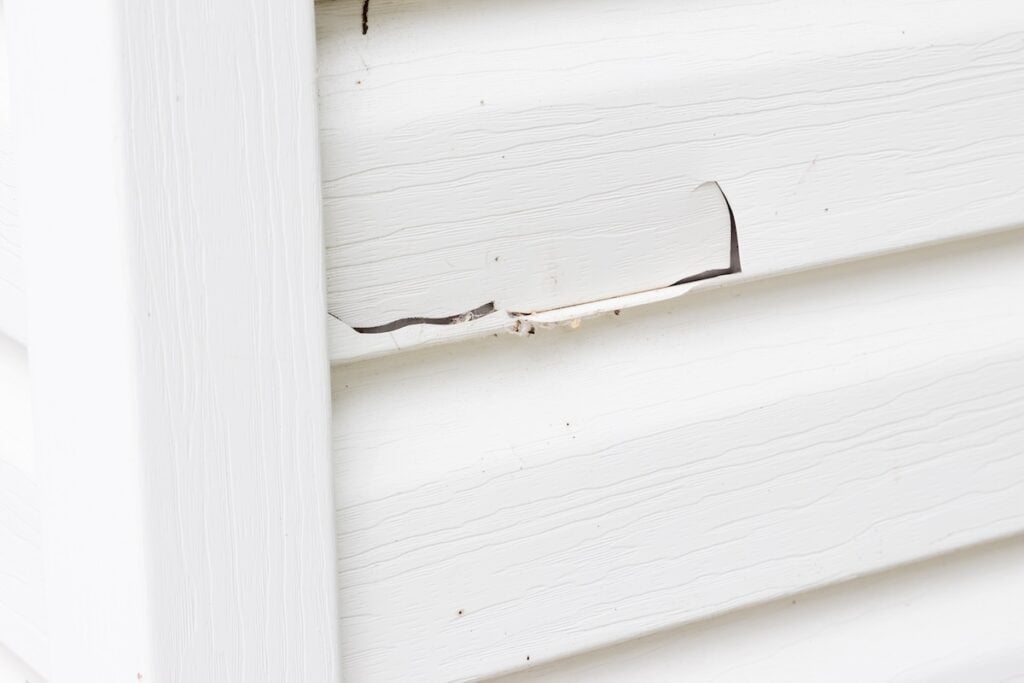 White vinyl siding on a house with a visible crack and hole in one panel near a corner, showing damage but no interior materials exposed
