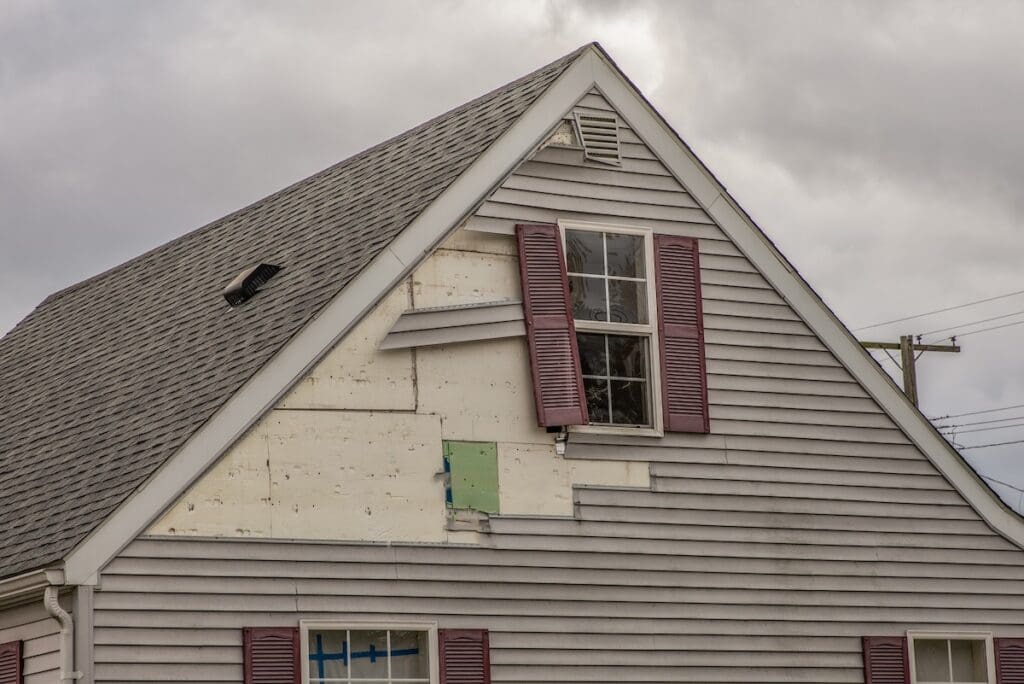 A house with mismatched siding and a boarded-up section near an upstairs window with closed burgundy shutters