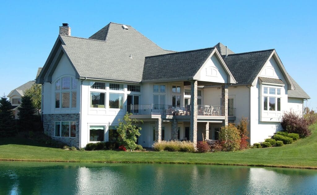 Large modern two-story house with gray roof, large windows, and a spacious balcony—perfect for relaxing while considering questions like “does homeowners insurance cover siding”—set by a pond with manicured lawn and colorful landscaping under a clear blue sky.