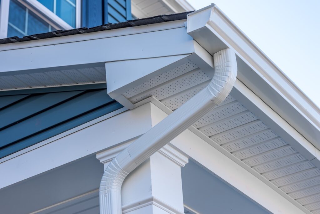 Close-up view of a house exterior showing a white rain gutter system attached to the roof edge near the soffit, with a downspout running beside blue siding and white trim. This highlights what is soffit on a house.