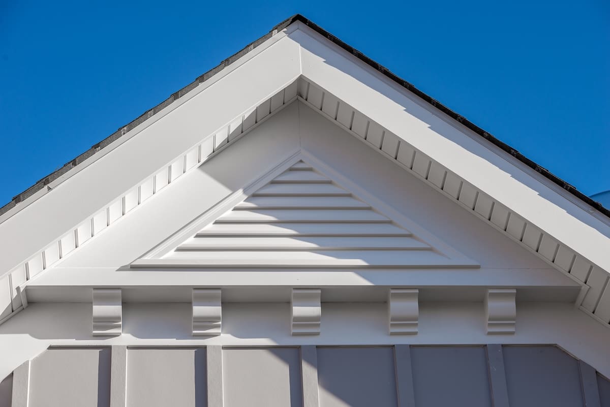 Close-up of a house gable with white trim, decorative molding, and a triangular vent with horizontal slats, showcasing details that help explain what is soffit on a house, set against a clear blue sky.