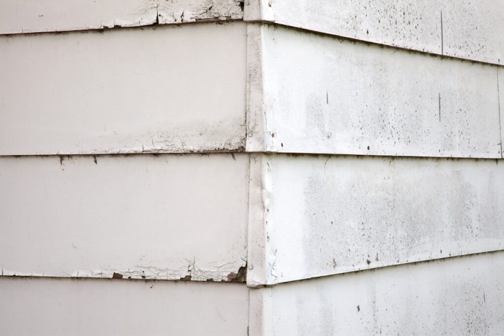 Close-up of the corner of a building with white, weathered siding. The paint is peeling and chipping, revealing signs of age