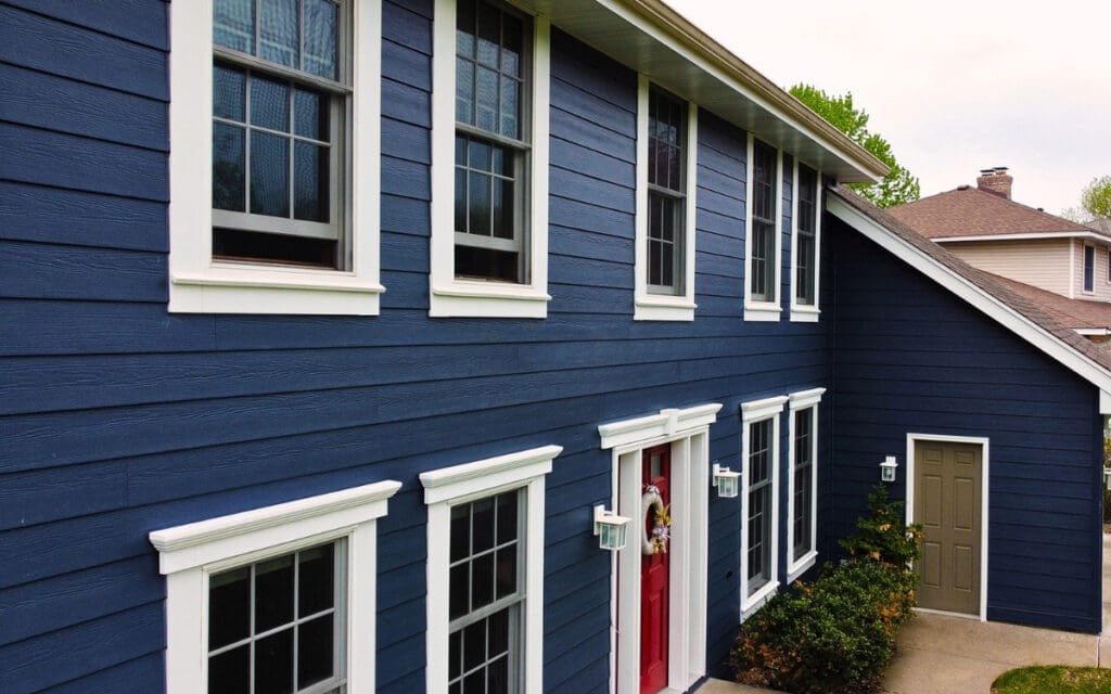 James Hardie siding cost Minnesota home with HardiePlank ColorPlus finish. A two-story house with James Hardie siding in Minnesota, blue exterior, white trim, multiple large windows, a red front door, and shrubs along the walkway leading to a side entrance.