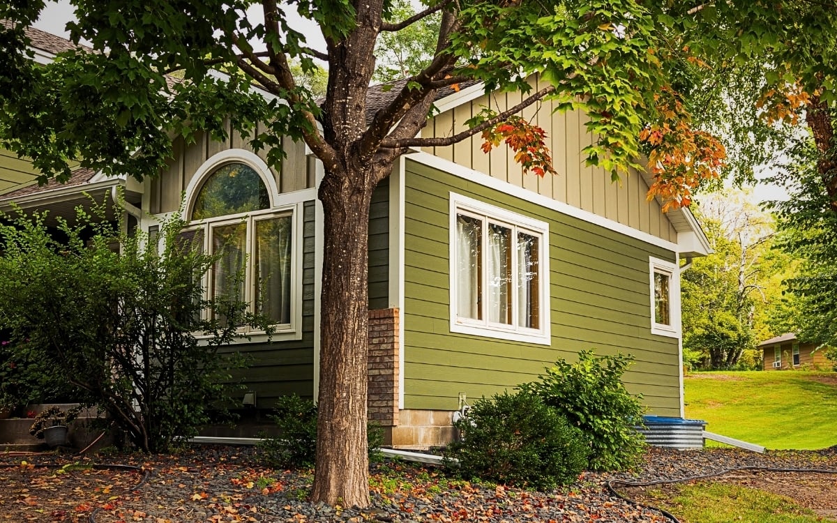 A green house with white trim, possibly showcasing hardie siding installation minnesota, sits among trees and shrubs. A lawn and small outbuilding are visible as some leaves suggest early autumn colors in the Twin Cities.