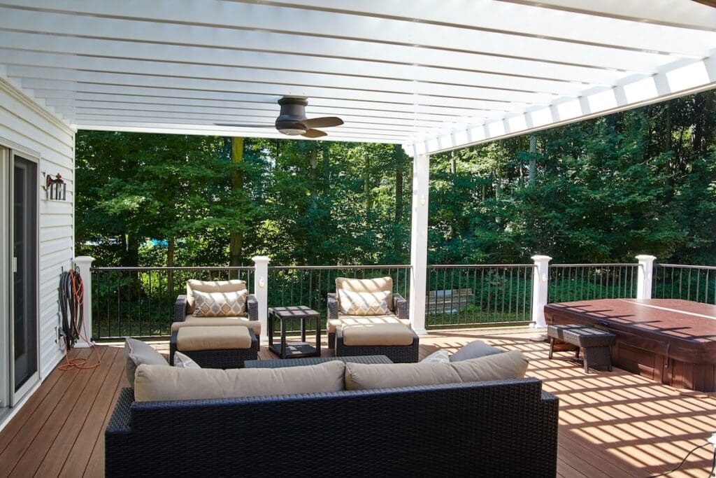 A deck with pergola features cushioned outdoor furniture, a ceiling fan, and a hot tub, overlooking a green, wooded backyard on a sunny day. Striped shadows from the pergola dance across the floor for a relaxing retreat.