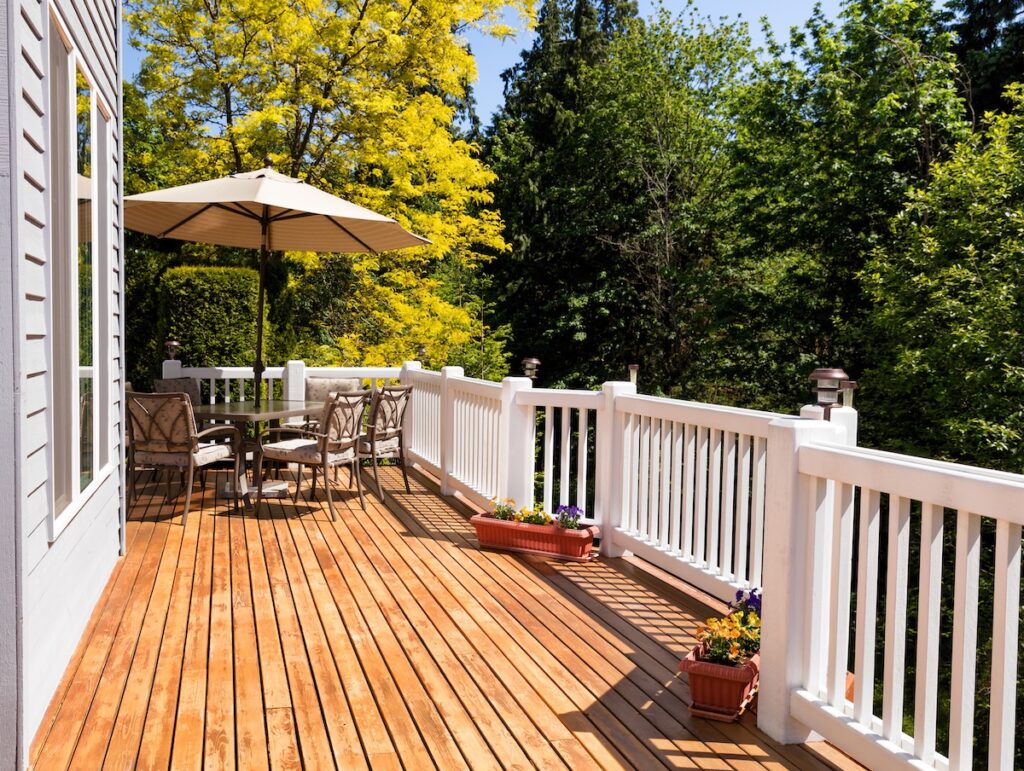 A wooden deck with white railings, one of many deck material options, features a patio table and chairs under a large beige umbrella. Flower boxes line the railing, while trees with green and yellow leaves surround the area.