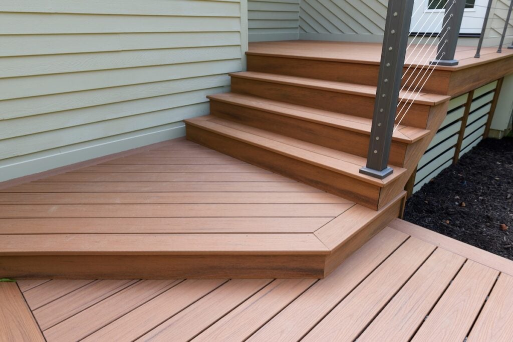 A modern outdoor wooden deck with steps leading up to a porch, showcasing clean lines and horizontal cable railing. Explore various deck material options next to light-colored siding and freshly mulched landscaping.