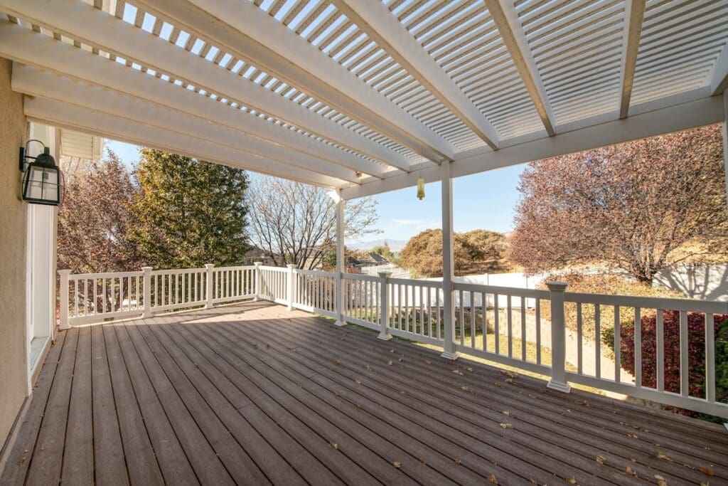 Terrace of a house with pergola roof and wood planks flooring. There is a white wooden handrailings and a view of trees and fence at the yard.