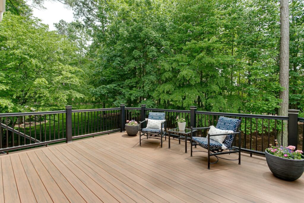 A spacious rooftop deck with two patterned chairs, a glass-top table, and potted plants, surrounded by a black railing and overlooking lush green trees.