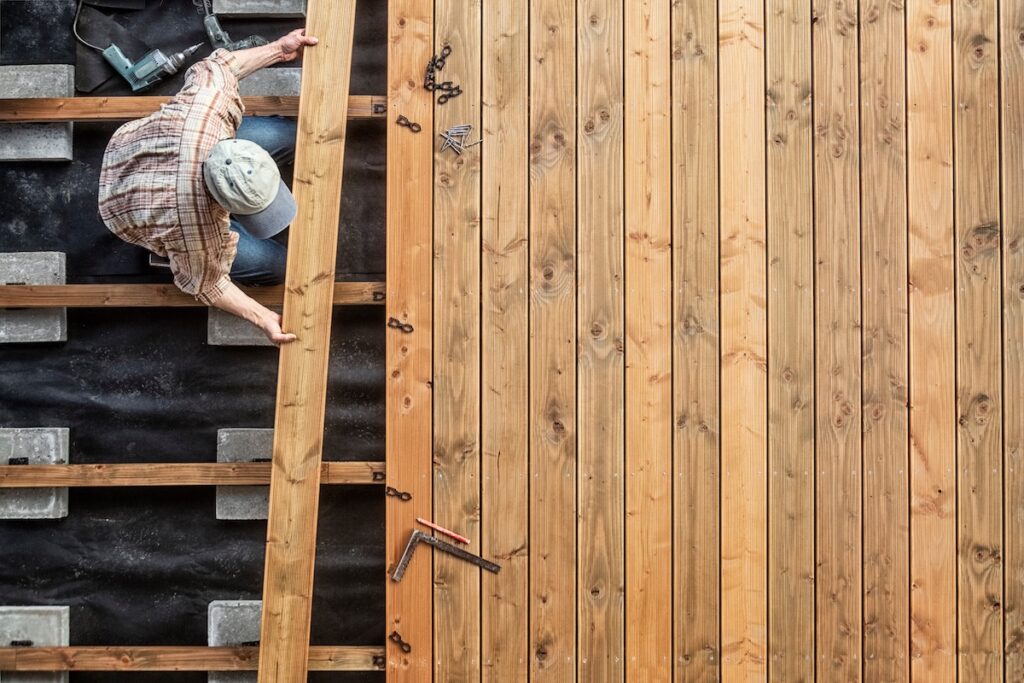 A person wearing a cap and plaid shirt installs wooden planks on a deck frame, using tools and screws, with an electric drill nearby. The right side showcases completed wooden decking, illustrating different deck material options.