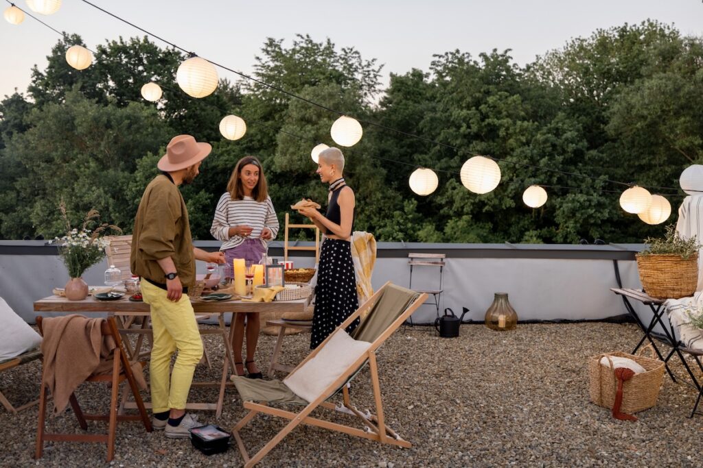 Three people stand around a wooden table on a rooftop deck decorated with string lights