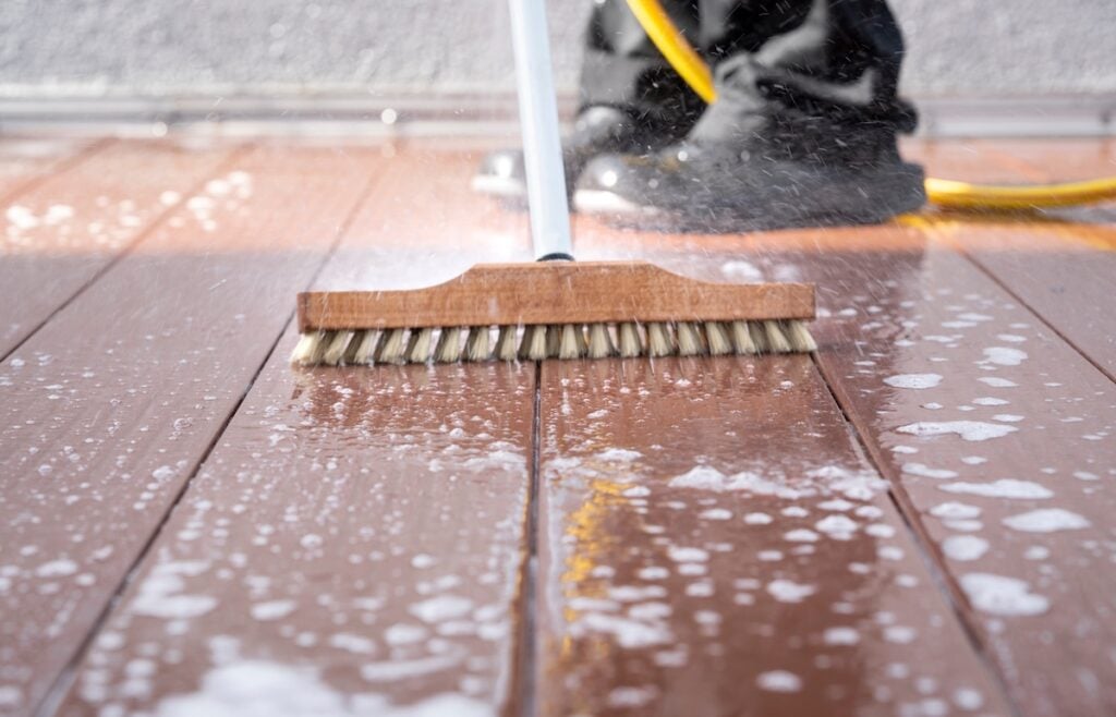 A close-up of a scrub brush at work on a soapy wooden deck highlights the deck cleaning process, with water and bubbles surrounding it. In the background, a person in black boots holds a hose, partially visible.