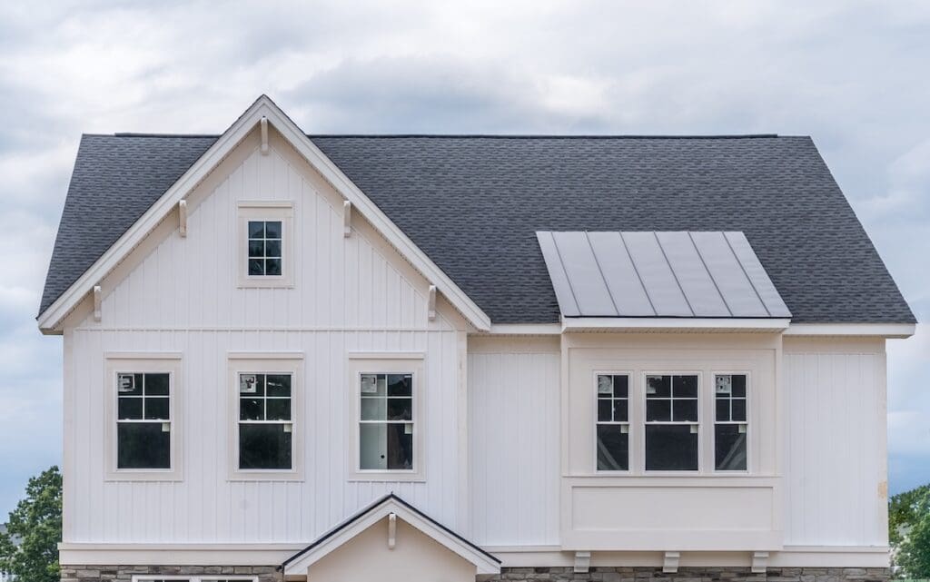 A two-story modern house with siding panels, black roof shingles, and several windows on the upper floor, set against a cloudy sky.