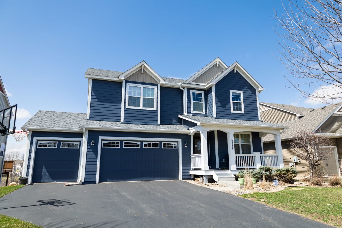 A two-story blue suburban house with white trim, a front porch, and a three-car garage sits on a paved driveway under a clear blue sky. There is some landscaping and a leafless tree in the yard.