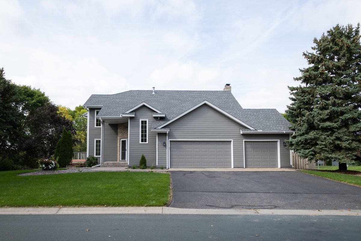 A modern two-story gray house with high-quality siding underlayment, a three-car garage, a well-kept lawn, and large trees on either side, viewed from the street on a cloudy day.