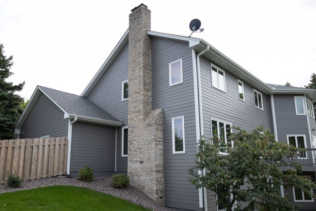 A modern two-story house with gray siding, white trim, a tall brick chimney, a satellite dish on the roof, and a fenced yard with green grass and shrubs.