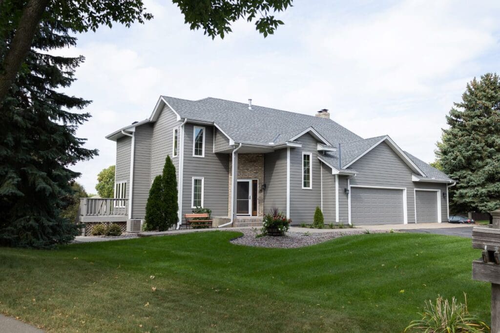 A two-story gray suburban house with white trim and quality siding underlayment, a large attached three-car garage, and well-kept lawn, surrounded by trees and shrubs on a cloudy day.