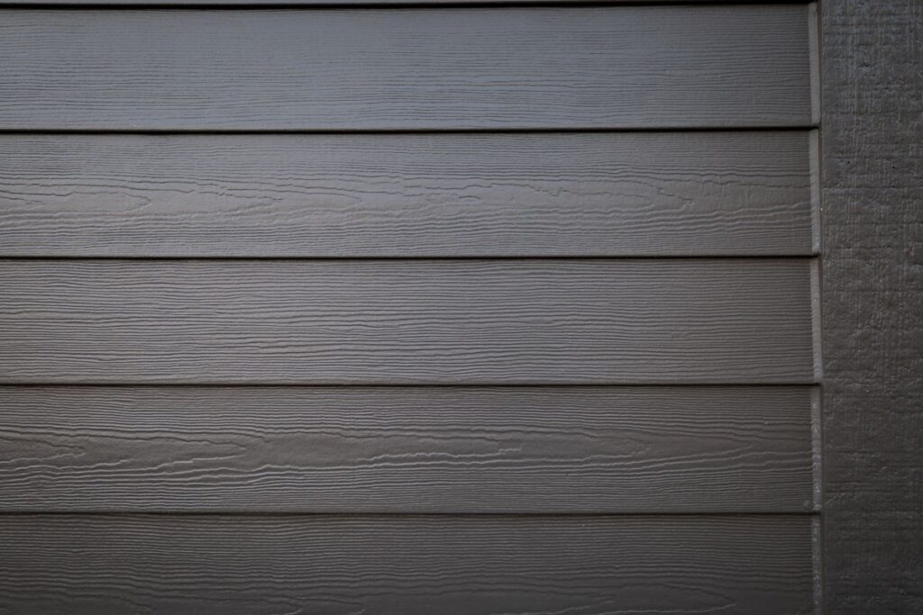 Close-up of a wall with horizontal, overlapping wooden or fiber cement siding panels. The surface has a subtle wood grain texture and is painted a neutral grey color. Light and shadow highlight the panel lines.