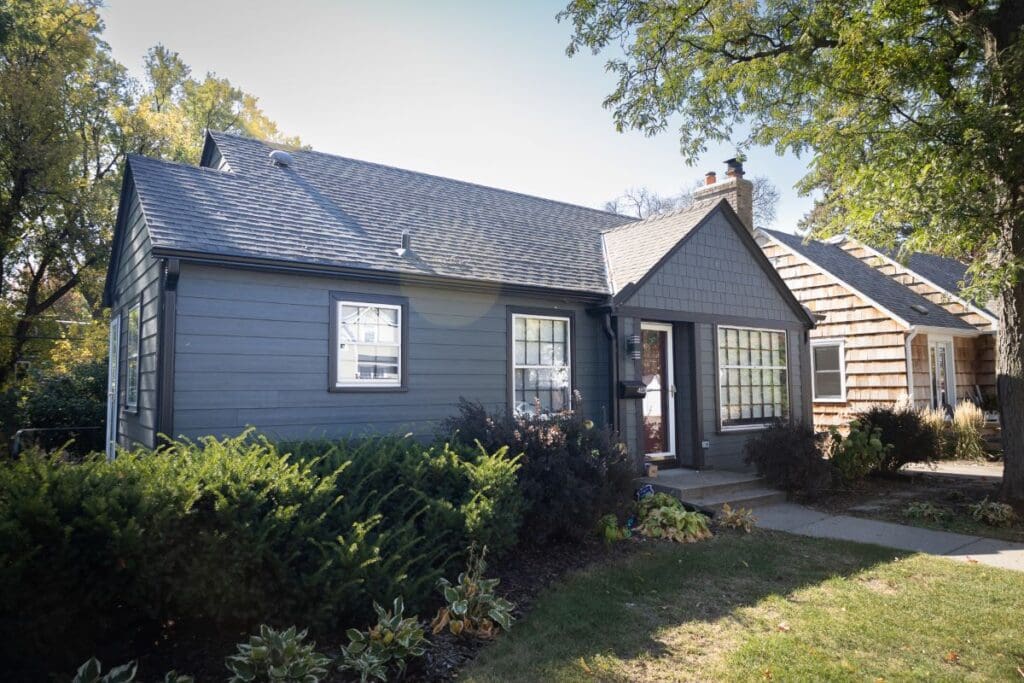 A small, gray cottage-style house with white-trimmed windows, quality siding underlayment, a shingled roof, and a front door with glass panels, surrounded by bushes and trees on a sunny day.
