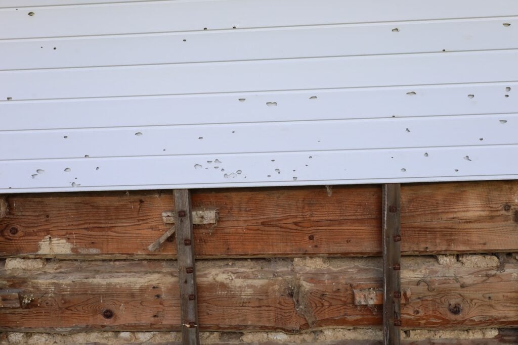 A white metal siding panel with multiple dents and holes, mounted above old, weathered wooden boards and exposed framing on a building wall