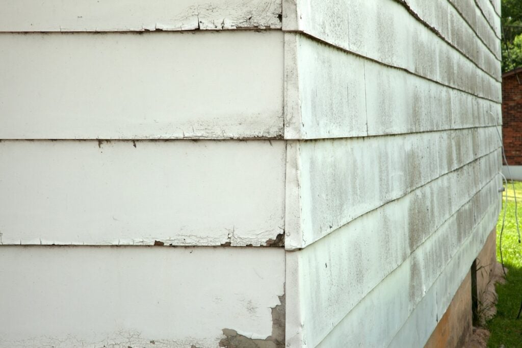 the corner of a weathered house with white, peeling paint on its wooden siding