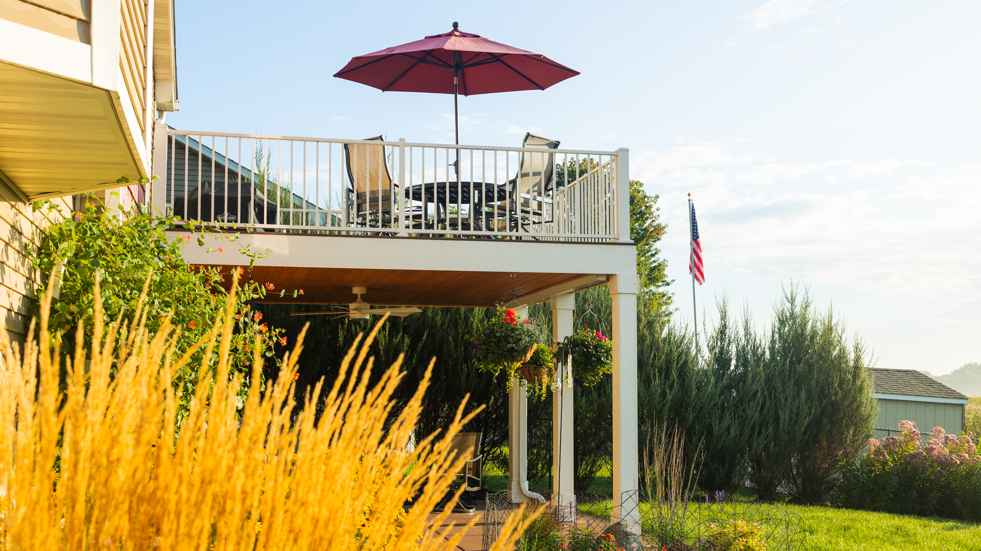 A raised deck with a white railing, outdoor table, chairs, and a red umbrella overlooks a garden with tall grasses, shrubs, hanging plants, and an American flag in the background, all lit by morning sunlight.