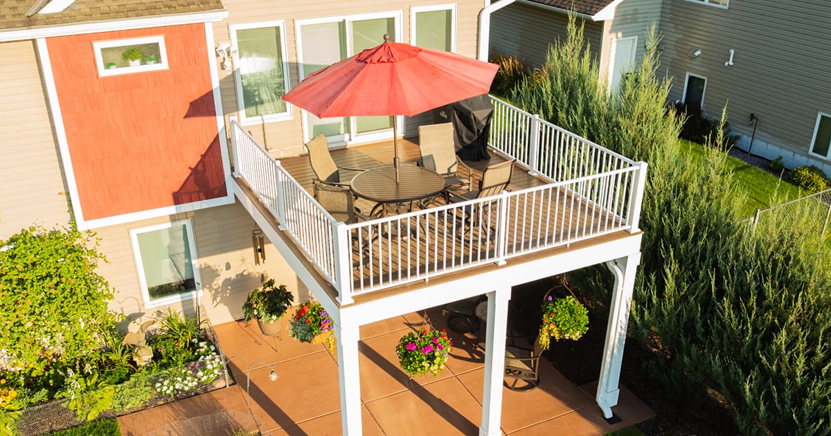 A raised backyard deck with white railings, a round table, four chairs, and a red umbrella, overlooking a patio with potted flowers and surrounded by greenery beside a tan house.
