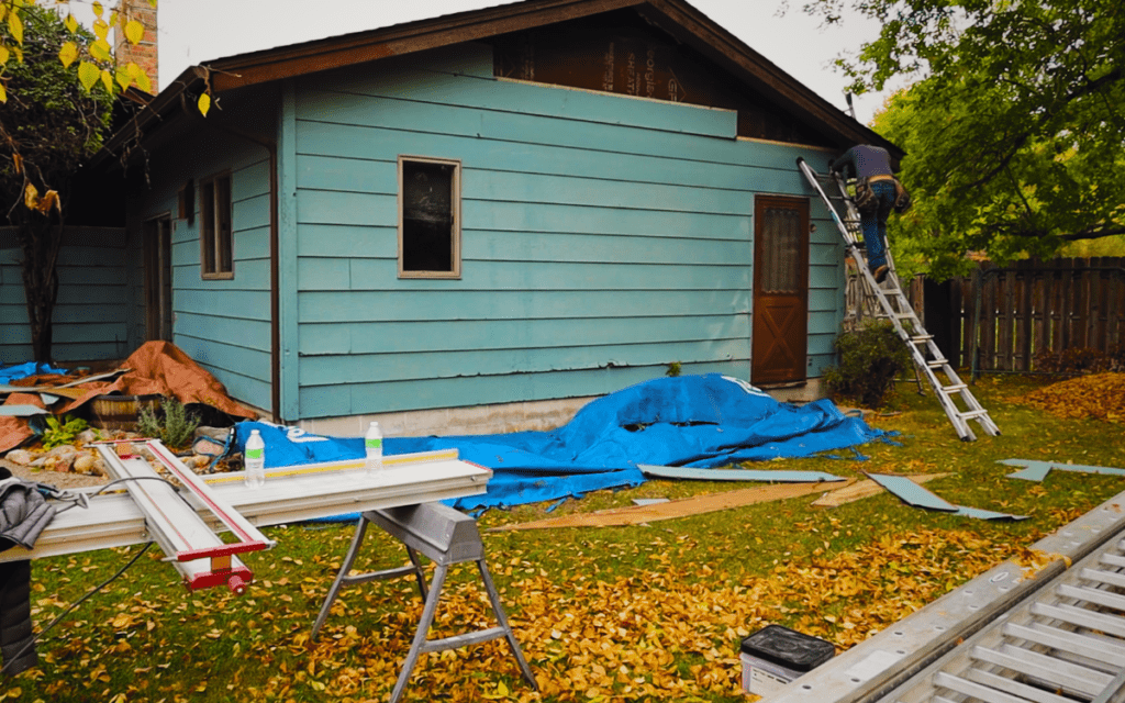 A person stands on a ladder working on the exterior wall of a blue house. Tools, boards, and a blue tarp—signs of siding damage repair by a Twin Cities siding contractor—are scattered among fallen autumn leaves.